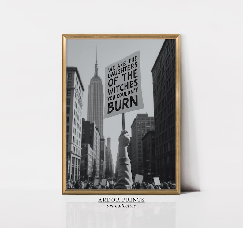 Może przedstawiać: Czarno-biała oprawiona grafika przedstawiająca scenę protestu w mieście z Empire State Building w tle. Ręka trzyma transparent z napisem "WE ARE THE DAUGHTERS OF THE WITCHES YOU COULDN'T BURN".