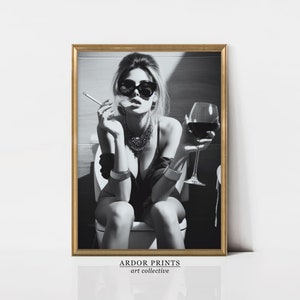 K&ouml;nnte beinhalten: Schwarzwei&szlig;foto einer Frau mit Sonnenbrille und schwarzem Oberteil, die auf einer Toilette sitzt und ein Glas Rotwein und eine Zigarette h&auml;lt.