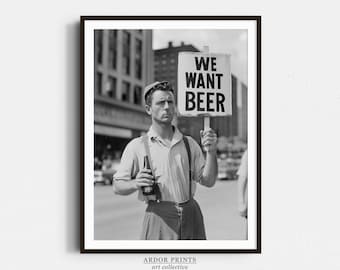 May include: Black and white photo of a man holding a sign that says "We Want Beer" and a bottle of beer. The man is wearing a white shirt and suspenders. The photo is in a black frame.