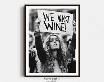 May include: Black and white photograph of a woman holding a sign that reads "We Want Wine!" The woman is wearing a denim jacket and has long, curly hair.