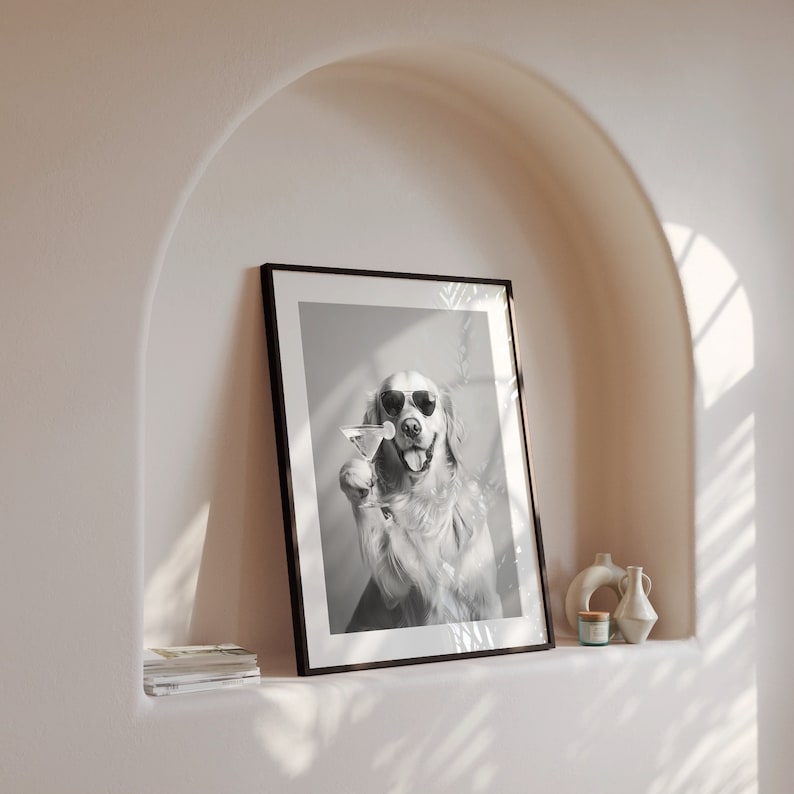 Op de afbeelding: Zwart-witfoto van een hond met een zonnebril die een martini-glas vasthoudt. De hond is een golden retriever en kijkt recht in de camera. De foto is ingelijst in een zwart frame en hangt aan een witte muur.
