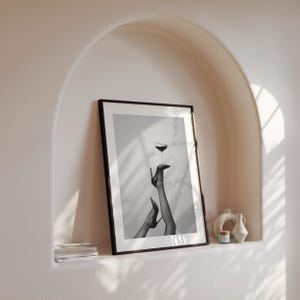 May include: Black and white framed art print featuring a glass of wine balanced on a high-heeled shoe. The image shows legs in stockings and heels, with a minimalist aesthetic. The artwork is displayed in a neutral-toned alcove.