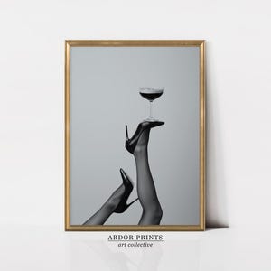 May include: Black and white framed art print featuring a pair of legs in black high heels, holding a coupe glass filled with a dark liquid. The print has a gold frame and the text "ARDOR PRINTS art collective" at the bottom.