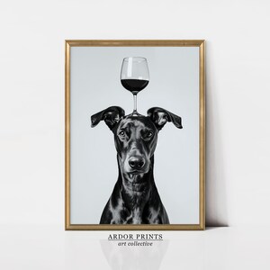 May include: Black and white photograph of a dog with a glass of red wine balanced on its head. The image is framed in a gold frame and has the text "ARDOR PRINTS art collective" at the bottom.
