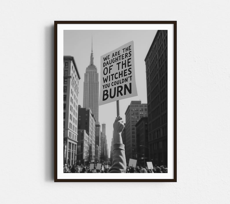 Może przedstawiać: Czarno-białe zdjęcie z protestu w mieście. Osoba trzyma transparent z napisem "WE ARE THE DAUGHTERS OF THE WITCHES YOU COULDN'T BURN", na tle Empire State Building.