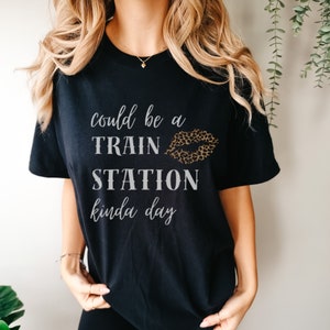 Op de afbeelding: Zwart T-shirt met de tekst "could be a TRAIN STATION kinda day" in witte en grijze letters. Een luipaardprint-lipdesign bevindt zich boven het woord "TRAIN".