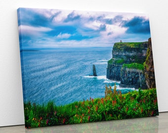 May include: A scenic view of the Cliffs of Moher in Ireland. The cliffs are tall and dramatic, with a green grassy top and a rugged, rocky face. The ocean is a deep blue, and the sky is a bright blue with white clouds. There are red flowers in the foreground.
