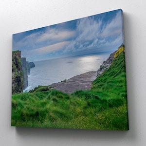 May include: A panoramic view of the rugged Irish coastline, featuring dramatic cliffs rising from the sea. Lush green grass covers the clifftops, while the ocean stretches out in the distance. The sky is a mix of blue and gray, with clouds casting shadows on the landscape.