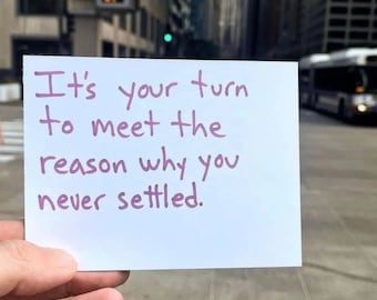 May include: A white card with purple text that reads, "It's your turn to meet the reason why you never settled." The card is held in front of a blurred city street scene with a bus.