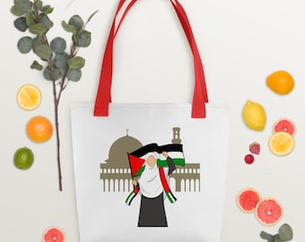 May include: White tote bag with red handles featuring a graphic of a person holding a Palestinian flag in front of a mosque silhouette. The flag has red, white, green, and black stripes. The person is wearing a hijab and a black dress.