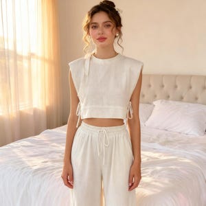 May include: A two-piece linen outfit in a cream color. The top is a sleeveless crop top with tie details on the sides and a tie at the neckline. The matching pants are high-waisted with a drawstring. The outfit is made of a lightweight fabric.