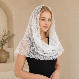 May include: A white lace head covering featuring a delicate floral pattern with scalloped edges. The fabric is semi-sheer and drapes over the head and shoulders, showcasing intricate needlework and a lightweight, airy texture throughout the design.