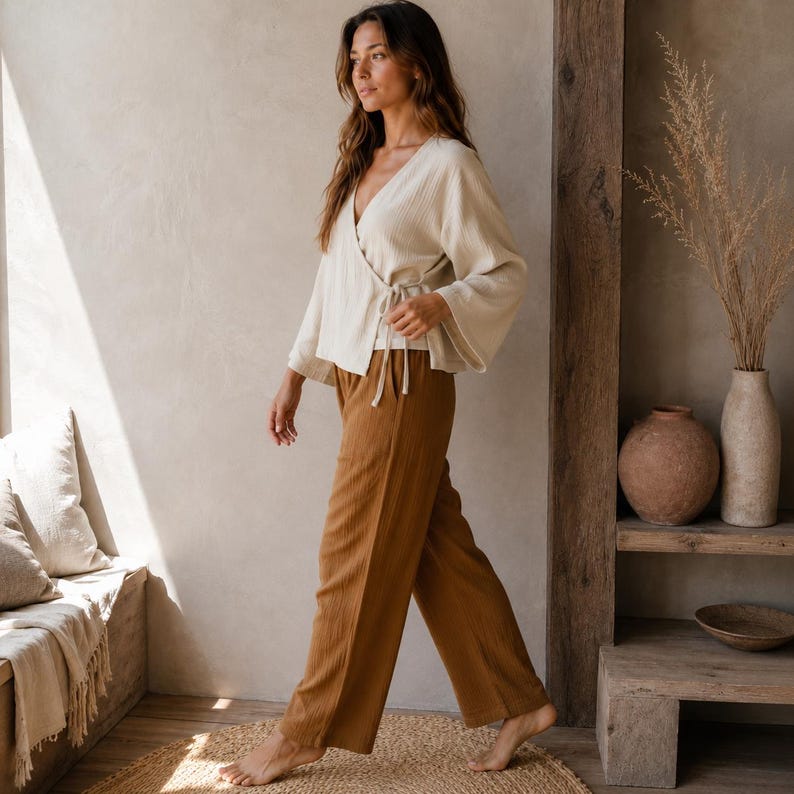 May include: A cream-colored wrap top with long, wide sleeves and a side tie closure, paired with loose-fitting, wide-leg trousers in a solid tobacco brown color. Both garments feature a subtle, vertical crinkled texture throughout the fabric.