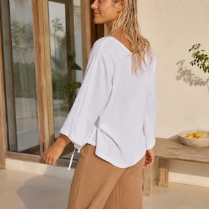 May include: A white long-sleeved top with a textured fabric, a wide neckline, and loose sleeves featuring thin ties at the cuffs. The garment is paired with tan trousers that have a visible textured weave and a relaxed, straight-leg fit.