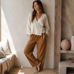 May include: A cream-colored long-sleeve wrap top with a side tie closure and a textured fabric finish, paired with loose-fitting, wide-leg trousers in a solid brown color. The garments feature a soft, crinkled material texture throughout.