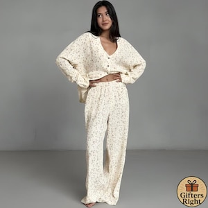 May include: Cream-colored pajama set with a floral pattern. The set includes a long-sleeved, button-down top and wide-leg pants. The fabric appears soft and lightweight, suitable for sleepwear or loungewear. The floral design features small, muted-color flowers.