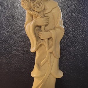 May include: A cream-colored ceramic figurine of a woman holding a bouquet of roses. The figurine is standing on a base with a floral design.