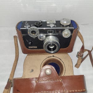 May include: A vintage black Argus C3 camera with a silver lens and controls, housed in a brown leather case. The camera has a rangefinder dial and a flash mount. The case is open, revealing a blue interior.