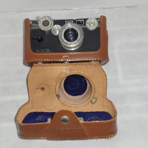 May include: A vintage black and silver camera with a leather case. The camera has a lens, dials, and a viewfinder. The brown leather case is open, revealing a blue interior. The camera is a classic piece of photography equipment.