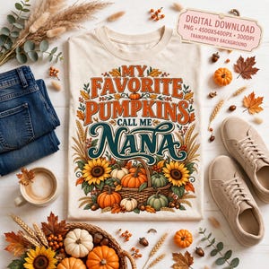 May include: A cream-colored t-shirt featuring a graphic with the text "MY FAVORITE PUMPKINS CALL ME NANA" surrounded by illustrations of pumpkins, sunflowers, and wheat. The design includes a woven basket filled with various pumpkins.