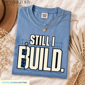 May include: A blue short-sleeved t-shirt with a crew neckline and a Comfort Colors label. The shirt features a graphic with the text STILL I BUILD in large cream-colored block letters, overlaid on a blue grid pattern with a crane illustration.
