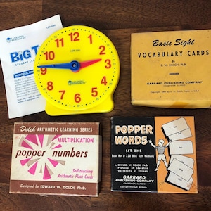May include: A collection of educational materials, including a yellow clock with red and blue hands, vocabulary cards, and flashcards for arithmetic and sight words. The clock has numbers 1-12. The flashcards are labeled "Popper Words" and "Multiplication".