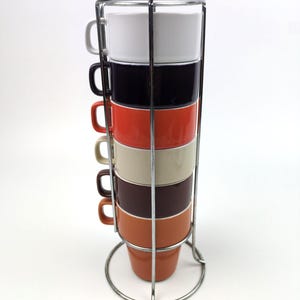 May include: A set of six stacked ceramic mugs in white, black, red, cream, dark brown, and terracotta, held within a vertical chrome wire rack. Each mug features a side handle and a smooth finish, organised in a tower formation within the metal frame.
