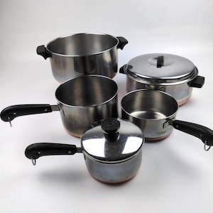 May include: A set of five stainless steel cooking pots and pans with copper-toned bases. The collection includes two lidded pots, two saucepans with long black handles, and one large stockpot with two side handles, all featuring black knobs.