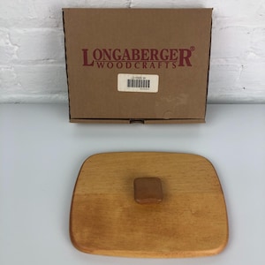 May include: A wooden lid for a Longaberger Woodcrafts storage container. The lid is light brown and has a small, round, wooden knob in the center.