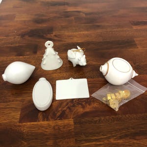 May include: A collection of white ceramic ornaments, including a teardrop shape, a Christmas tree, a pig with wings, an oval, a rectangular plaque, and a round ornament with gold accents. A small bag of gold-colored hooks is also included.