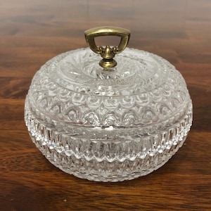 May include: A clear, round glass jar with a decorative lid and a brass handle. The jar has a detailed, textured pattern. The lid is also clear and has a similar design. The handle is a curved, brass-colored metal.