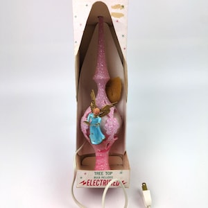 以下が含まれることがあります： 「Christmas Decorations by Decor」と書かれた箱に入った、ピンク色のグリッター付きクリスマスツリートップ。中央の球体には本を持った青いローブの天使のフィギュアがあり、その上に尖塔が付いています。電気コードが含まれています。