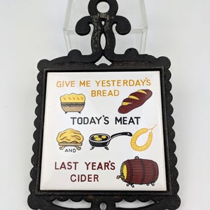 May include: A black cast iron trivet with a decorative scrollwork frame holding a square ceramic tile. The tile features illustrations of bread, meat, and a barrel, with the text: GIVE ME YESTERDAY'S BREAD, TODAY'S MEAT, AND LAST YEAR'S CIDER.
