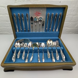 May include: A silver-toned flatware set housed in a wooden storage case with a blue velvet interior. The set includes various spoons, forks, and knives, with a label reading From the Holmes & Edwards Collection. The utensils feature decorative handles.