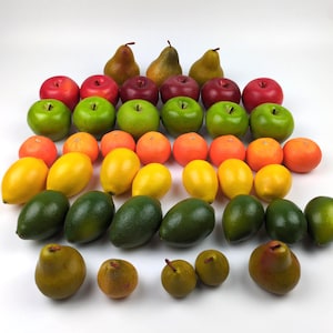 May include: A collection of artificial fruit including red and green apples, orange citrus fruits, yellow lemons, dark green avocados, and brown pears. The items are arranged in rows on a white surface, showcasing a variety of colors and shapes.