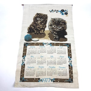 May include: A beige linen wall calendar for the year 1977 featuring two fluffy dark brown kittens and a blue ball of yarn. The calendar displays all twelve months in a grid, framed by a brown border decorated with small blue and black flowers.
