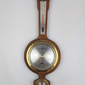 May include: A wall-mounted weather station featuring a wooden frame with a central barometer, a vertical thermometer, and a smaller circular gauge at the bottom. The barometer dial displays the text RAIN, CHANGE, FAIR, and DRY with a brass bezel.