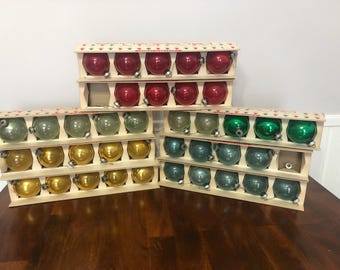 May include: A collection of round glass ornaments arranged in cardboard display boxes. The set includes red, gold, light green, dark green, and teal spheres, each featuring a silver-colored metal cap at the top for hanging purposes.