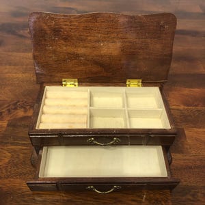 May include: A vintage wooden jewelry box with a hinged lid and a pull-out drawer. The interior features compartments and ring rolls lined with a cream-colored fabric. The box has brass hinges and drawer pulls, and is made of dark brown wood.