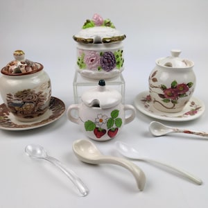 May include: A collection of three ceramic lidded jars with matching saucers, featuring floral and fruit motifs, accompanied by three small spoons. The set includes a brown landscape pattern, a pink and purple floral design, and a strawberry print.