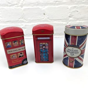 May include: Three decorative metal tea tins. The first is a red double-decker bus with "NEW ENGLISH TEAS" and "LONDON TRANSPORT" text. The second is a red telephone box labeled "ENGLISH AFTERNOON TEA". The third is a cylindrical Union Jack tin.
