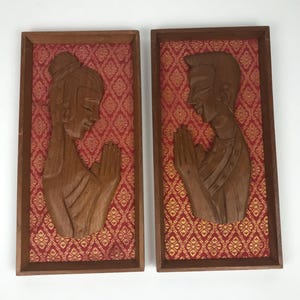 Pode incluir: Duas placas de parede retangulares de madeira com figuras esculpidas em posição de oração. Cada figura é montada sobre um fundo de tecido vermelho com um padrão geométrico dourado. As molduras são feitas de madeira castanho-escura.