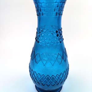 May include: A vibrant blue glass vase featuring a flared rim and a tapered neck. The body is decorated with a textured pattern of raised squares near the top, a band of small circular dots in the middle, and a diamond lattice design at the base.