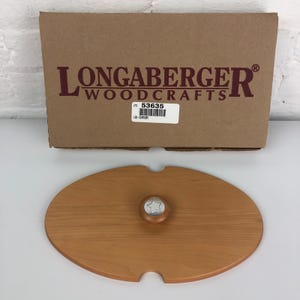 May include: A wooden Longaberger Woodcrafts oval-shaped lid with a star-shaped knob. The lid has a brown stain and is labeled with the item number "53635".