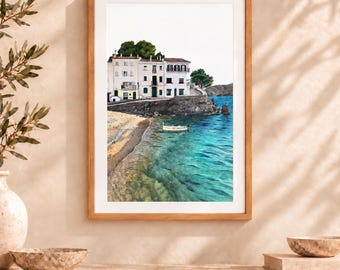May include: A framed art print featuring a coastal scene with white multi-story buildings perched on a rocky cliff above a sandy beach and turquoise water. A small white boat floats near the shore, and green trees are visible on the hillside.