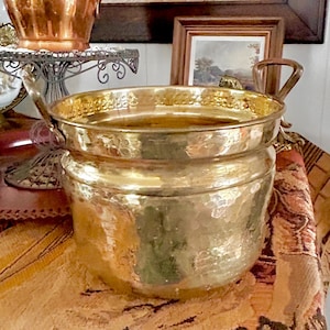May include: A hammered brass planter with two handles sits on a patterned textile. A copper pitcher with purple flowers and a decorative lamp are in the background. A framed picture of a landscape is also visible.