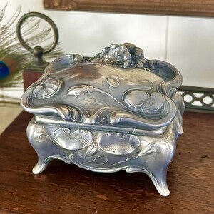 Könnte beinhalten: Eine silberfarbene Schmuckschatulle mit einem verzierten floralen Design auf Deckel und Seiten. Die Schatulle hat geschwungene Kanten und steht auf kleinen, dekorativen Füßen. Die Schatulle ist aus Metall und hat eine Vintage-Ästhetik.