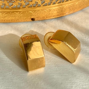 May include: A pair of gold-colored, geometric clip-on earrings. The earrings have a rectangular shape with a slightly curved design. The background includes a gold-colored, ornate tray with a floral design.