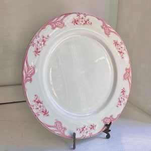 以下が含まれることがあります： 縁にピンクの花柄が施された白いセラミック製のプレート。プレートは金属製のスタンドの上に置かれています。
