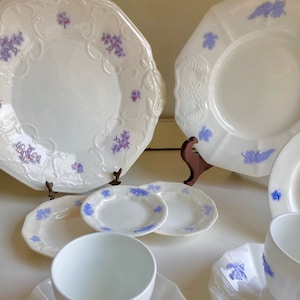 May include: A set of white porcelain plates and cups featuring a delicate blue floral design. The set includes a large plate, a smaller plate, a saucer, and an egg cup.